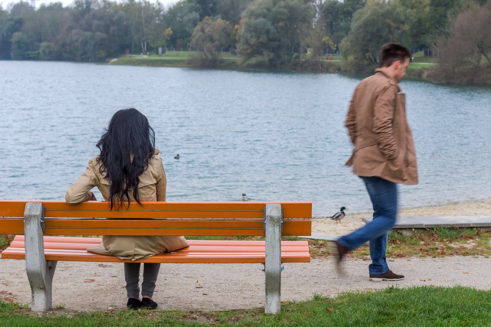 Frau sitzt am See alleine auf einer Bank. Mann geht weg